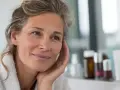 Une femme d'âge mûr, aux cheveux gris et portant une robe de chambre, se regarde dans le miroir. Elle a un sourire doux, comme si elle pensait à un trèfle rouge porte-bonheur.