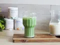 Un verre de matcha latte frais, avec des pots de poudre de matcha et une carafe de lait végétal en arrière-plan.