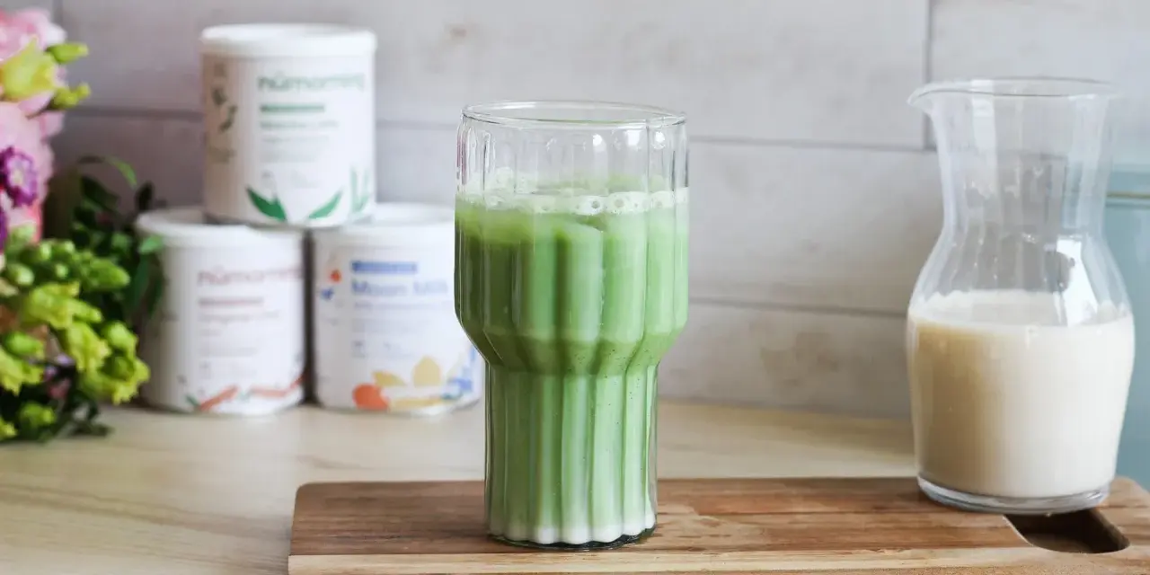 Un verre de matcha latte frais, avec des pots de poudre de matcha et une carafe de lait végétal en arrière-plan.