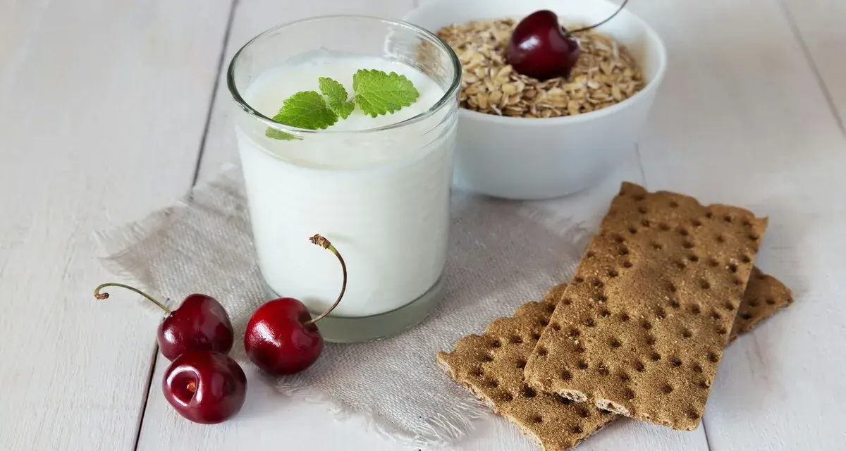Un verre de yaourt lait végétal, garni de menthe, avec des cerises et des crackers sur une table en bois.