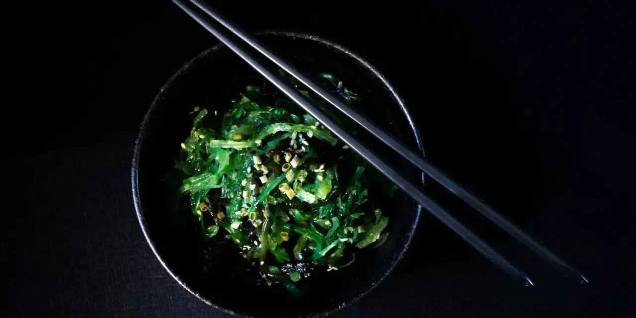 Salade de wakame algue, garnie de graines de sésame, dans un bol noir avec des baguettes.