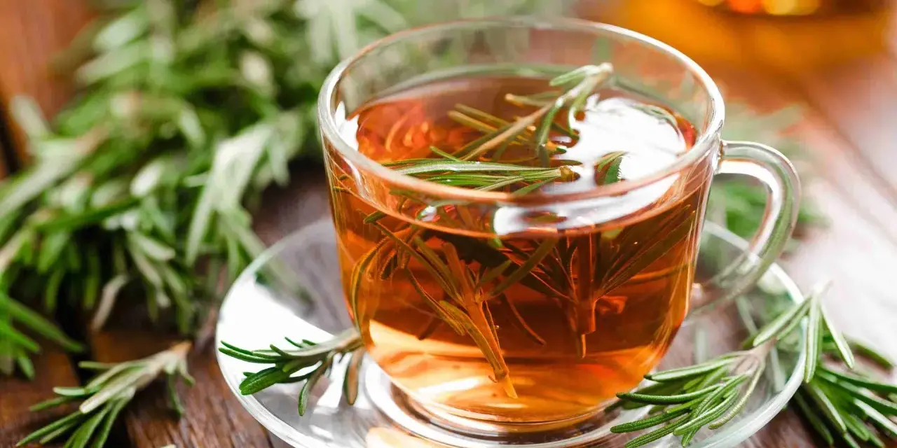 Une tasse de tisane de romarin fumante, garnie de brins frais, sur une soucoupe en verre, entourée de romarin sur fond de bois.