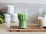 Un verre de matcha latte frais, avec des pots de poudre de matcha et une carafe de lait végétal en arrière-plan.