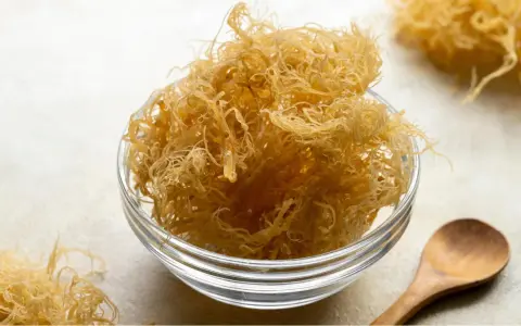 Un bol en verre rempli de **irish moss** séché, ressemblant à des fils fins et dorés. Une cuillère en bois repose à côté.