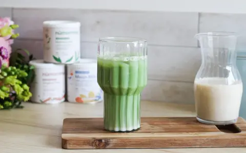 Un verre de matcha latte frais, avec des pots de poudre de matcha et une carafe de lait végétal en arrière-plan.