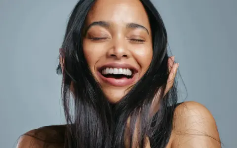 Une femme rit aux éclats, ses yeux fermés. Ses cheveux noirs encadrent son visage radieux, témoignant de la beauté que la vitamine B7 peut apporter.