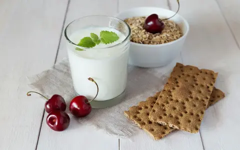 Un verre de yaourt lait végétal, garni de menthe, avec des cerises et des crackers sur une table en bois.