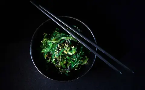 Salade de wakame algue, garnie de graines de sésame, dans un bol noir avec des baguettes.
