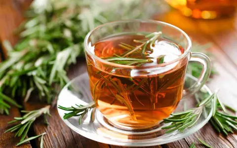 Une tasse de tisane de romarin fumante, garnie de brins frais, sur une soucoupe en verre, entourée de romarin sur fond de bois.
