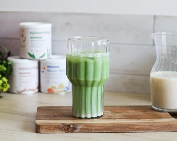 Un verre de matcha latte frais, avec des pots de poudre de matcha et une carafe de lait végétal en arrière-plan.