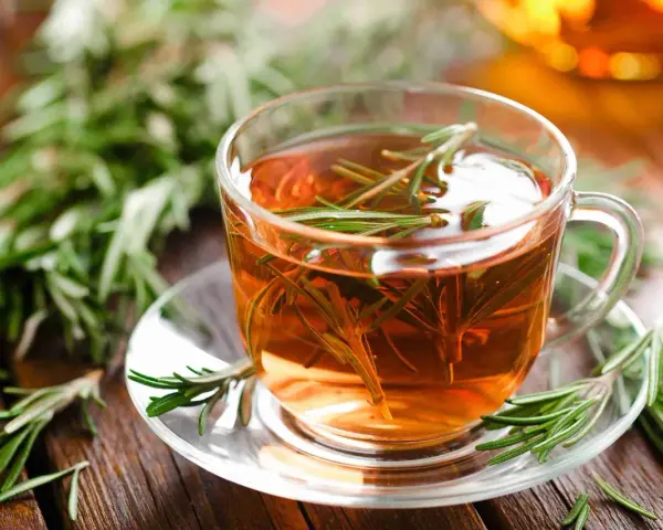 Une tasse de tisane de romarin fumante, garnie de brins frais, sur une soucoupe en verre, entourée de romarin sur fond de bois.
