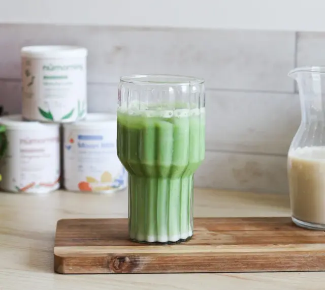 Un verre de matcha latte frais, avec des pots de poudre de matcha et une carafe de lait végétal en arrière-plan.