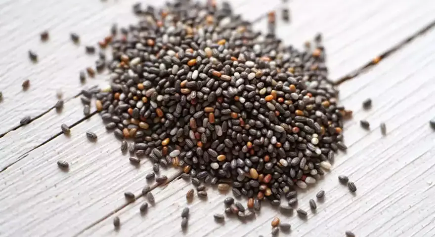 Un tas de graine de chia germé, petites et noires, sur un fond de bois blanc.