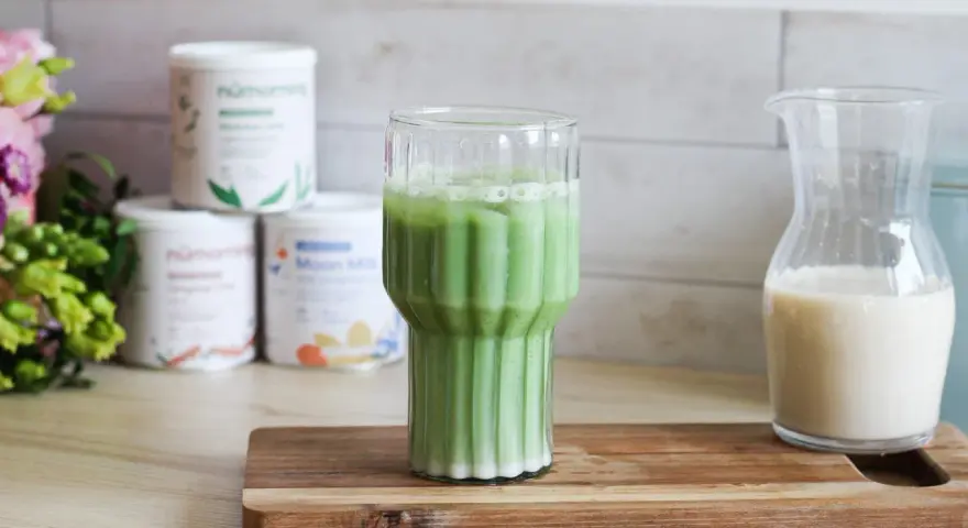 Un verre de matcha latte frais, avec des pots de poudre de matcha et une carafe de lait végétal en arrière-plan.