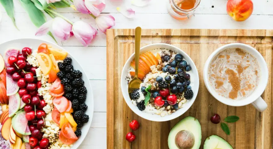 Un bol de yaourt aux fruits rouges, myrtilles et amandes, un plateau de fruits frais, un avocat et une tasse de café. Un repas sain, un aliment anti inflammatoire.