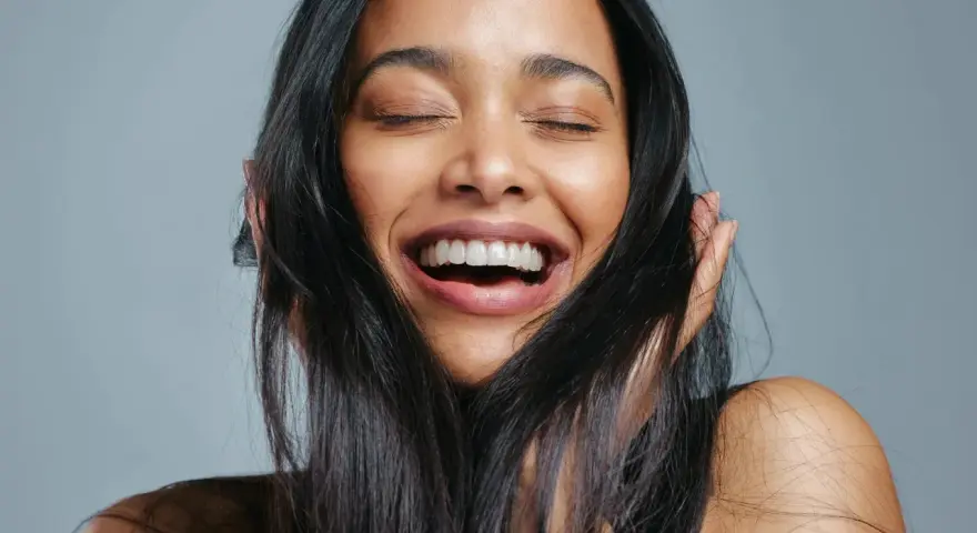 Une femme rit aux éclats, ses yeux fermés. Ses cheveux noirs encadrent son visage radieux, témoignant de la beauté que la vitamine B7 peut apporter.
