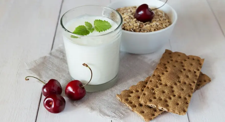 Un verre de yaourt lait végétal, garni de menthe, avec des cerises et des crackers sur une table en bois.
