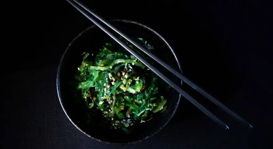 Salade de wakame algue, garnie de graines de sésame, dans un bol noir avec des baguettes.