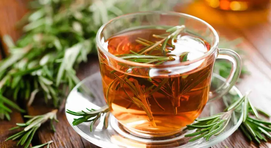 Une tasse de tisane de romarin fumante, garnie de brins frais, sur une soucoupe en verre, entourée de romarin sur fond de bois.