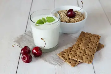 Un verre de yaourt lait végétal, garni de menthe, avec des cerises et des crackers sur une table en bois.