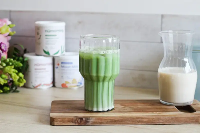 Un verre de matcha latte frais, avec des pots de poudre de matcha et une carafe de lait végétal en arrière-plan.